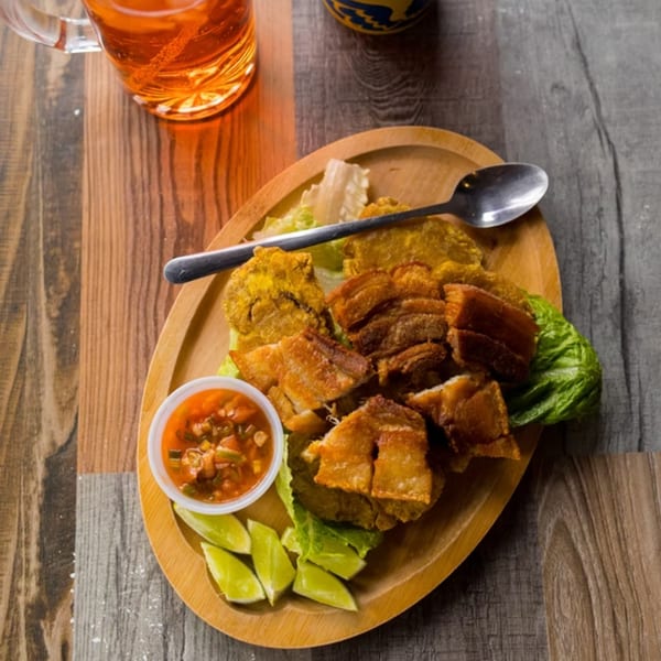 A wooden plate of Colombian chicharrón, tostones, salsa, and lime wedges, with a drink.