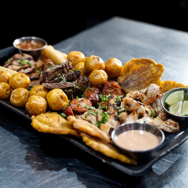 A large platter of Colombian food, including grilled meats, potatoes, plantains, and dipping sauces.