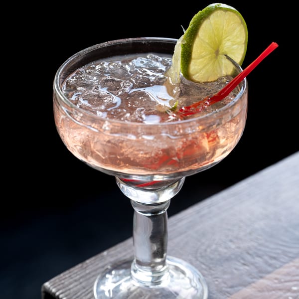 A pink cocktail with ice, a lime wedge, and a red straw in a margarita glass.