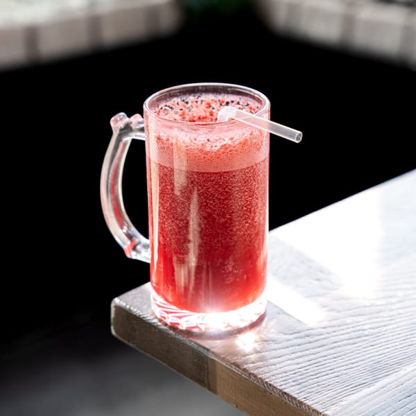 A vibrant red Colombian fruit juice in a glass mug with a straw on a sunlit wooden table.