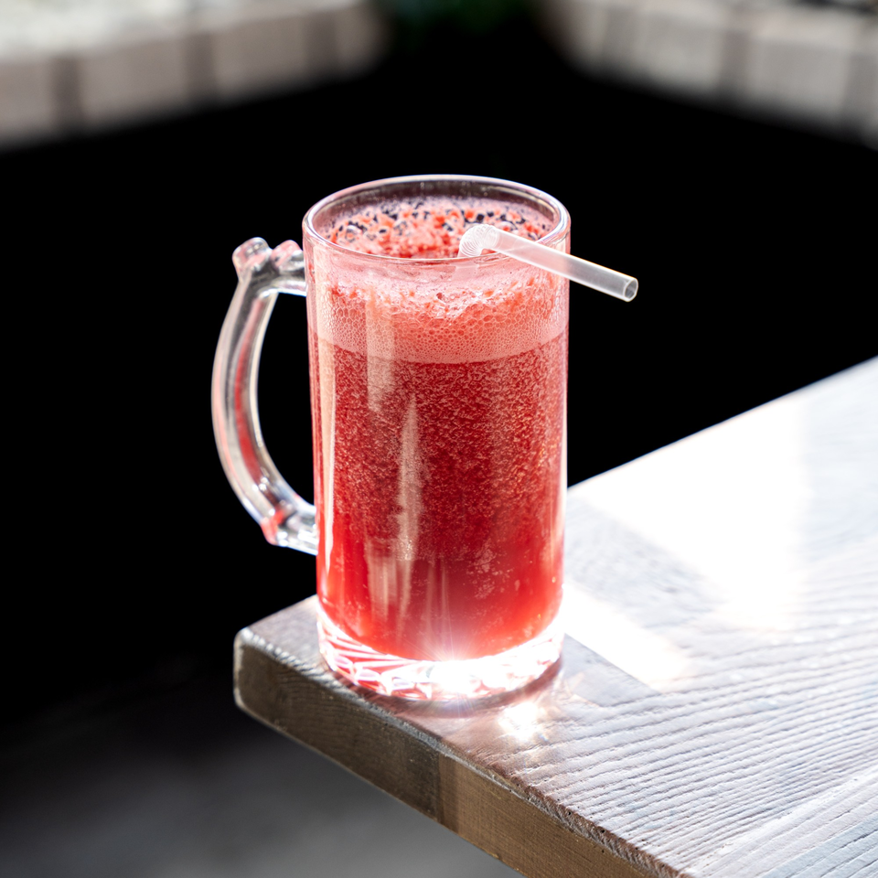 A vibrant red Colombian fruit juice in a glass mug with a straw on a sunlit wooden table.