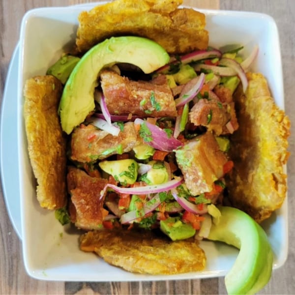 A white bowl of Chapa Paisa chicharron, avocado, red onion, and tomato salad with fried plantains.