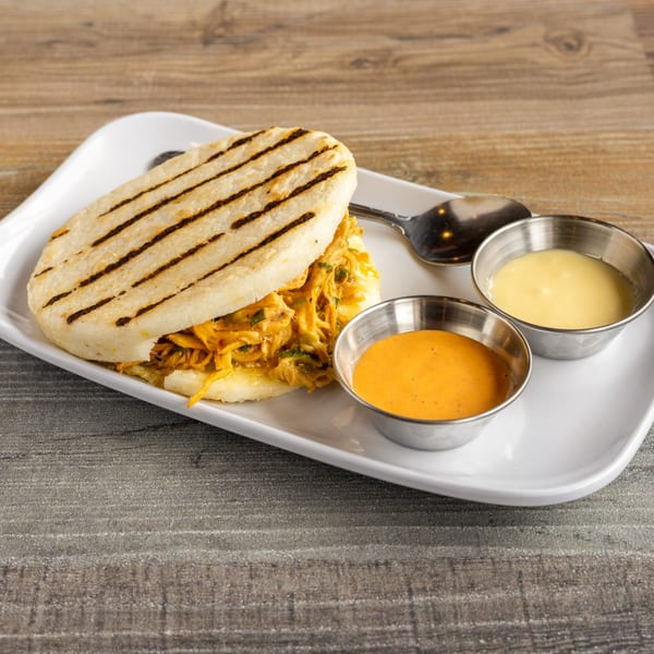 A Colombian arepa filled with shredded chicken, served with two dipping sauces on a white plate.