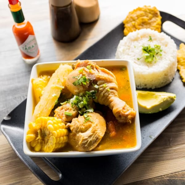 Colombian chicken sancocho with corn, yucca, rice, avocado, and plantains on a black tray.